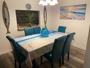 A dining area with a table covered by a beach-themed tablecloth, surrounded by blue chairs, a blue vase, and a painting of a beach scene on the wall.