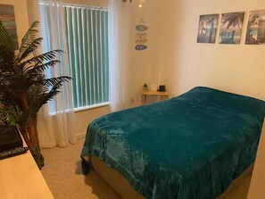 A bedroom with a bed, a potted plant, a window with blinds, a small table with a lamp, and wall art.