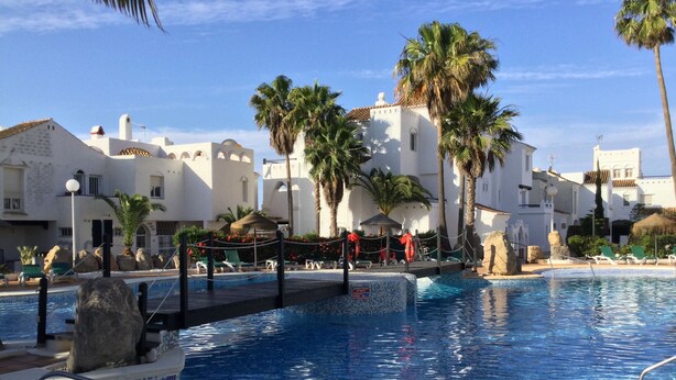 A pool area with clear blue water, surrounded by white buildings with arched windows and palm trees.