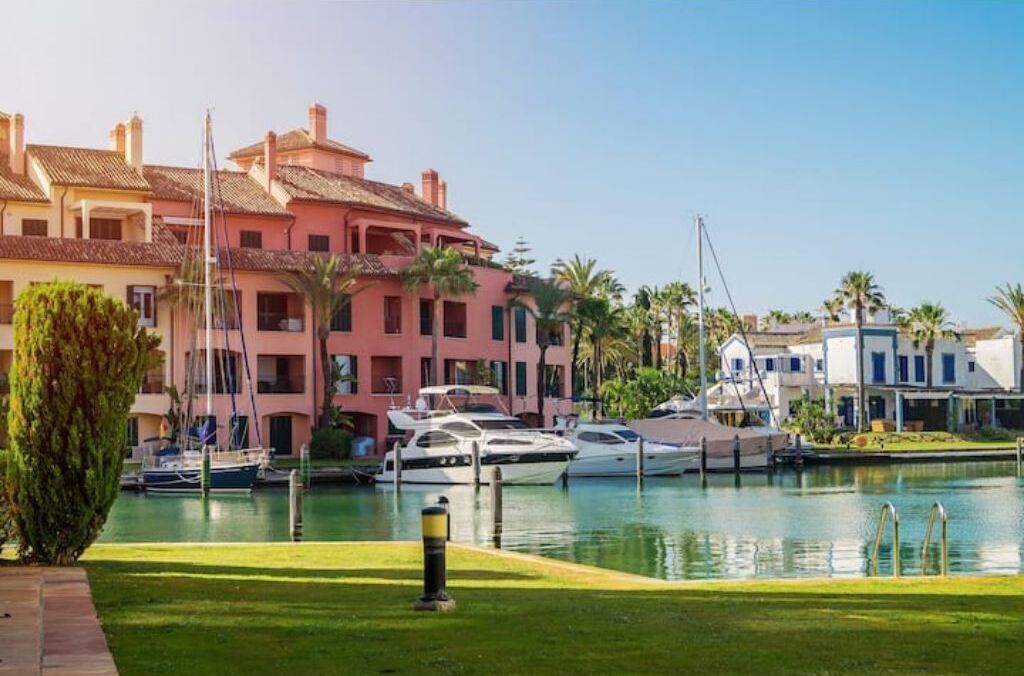 Un puerto deportivo con barcos amarrados, rodeado de edificios con balcones y palmeras.