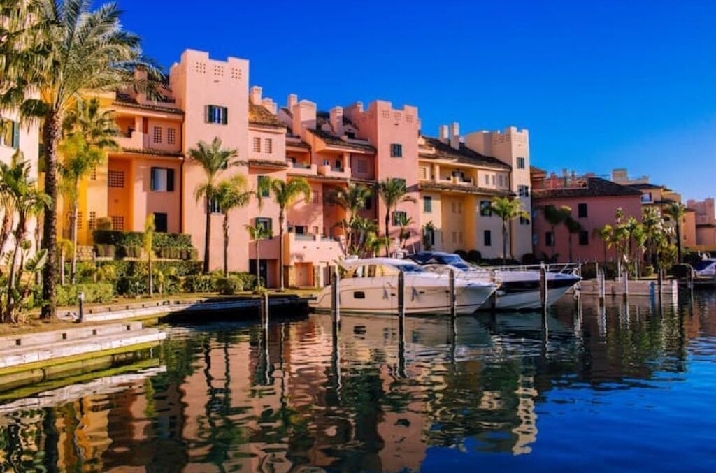 Un puerto deportivo con barcos amarrados, rodeado de edificios de varios pisos con balcones, palmeras y un cielo azul despejado.