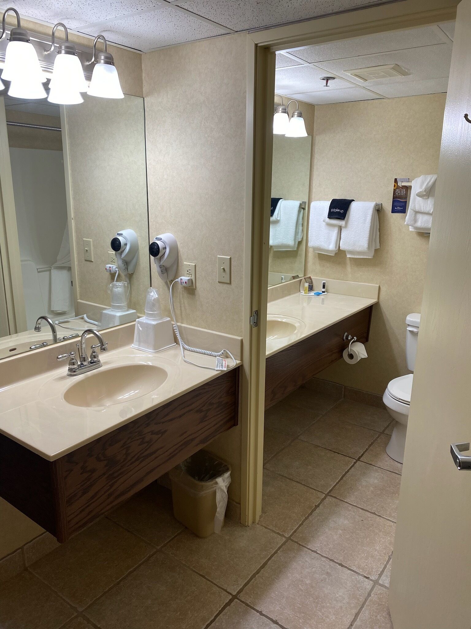 A modern bathroom with a large mirror, two sinks, a hairdryer, and a toilet.