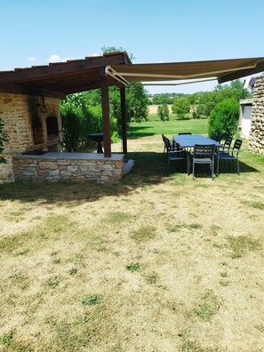 A stone-built outdoor kitchen with a pergola, a table with chairs, and a view of a grassy field.