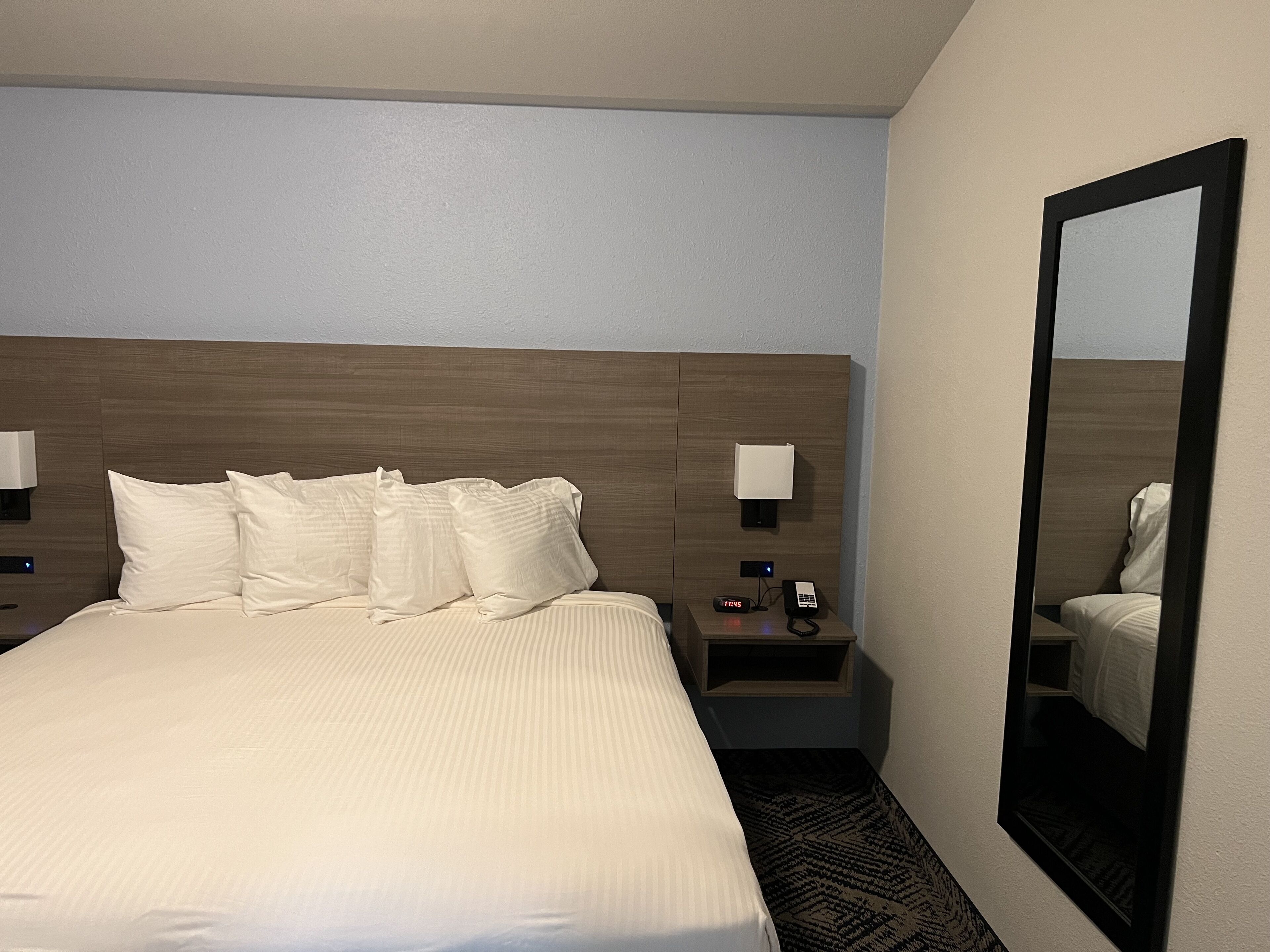 Quarto de hotel com uma cama grande, duas mesas de cabeceira, um criado-mudo, um espelho e uma luminária fixada na parede.