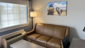 A room with a brown leather sofa, a table, a lamp, a large window, and a painting of a train on the wall.