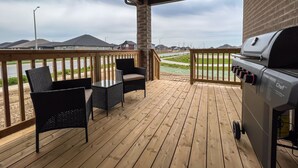 A covered outdoor deck with a grill, wicker chairs, and a view of a residential area. - GLOBALSTAY Brand New House in Port Elgin (Saugeen Shores)