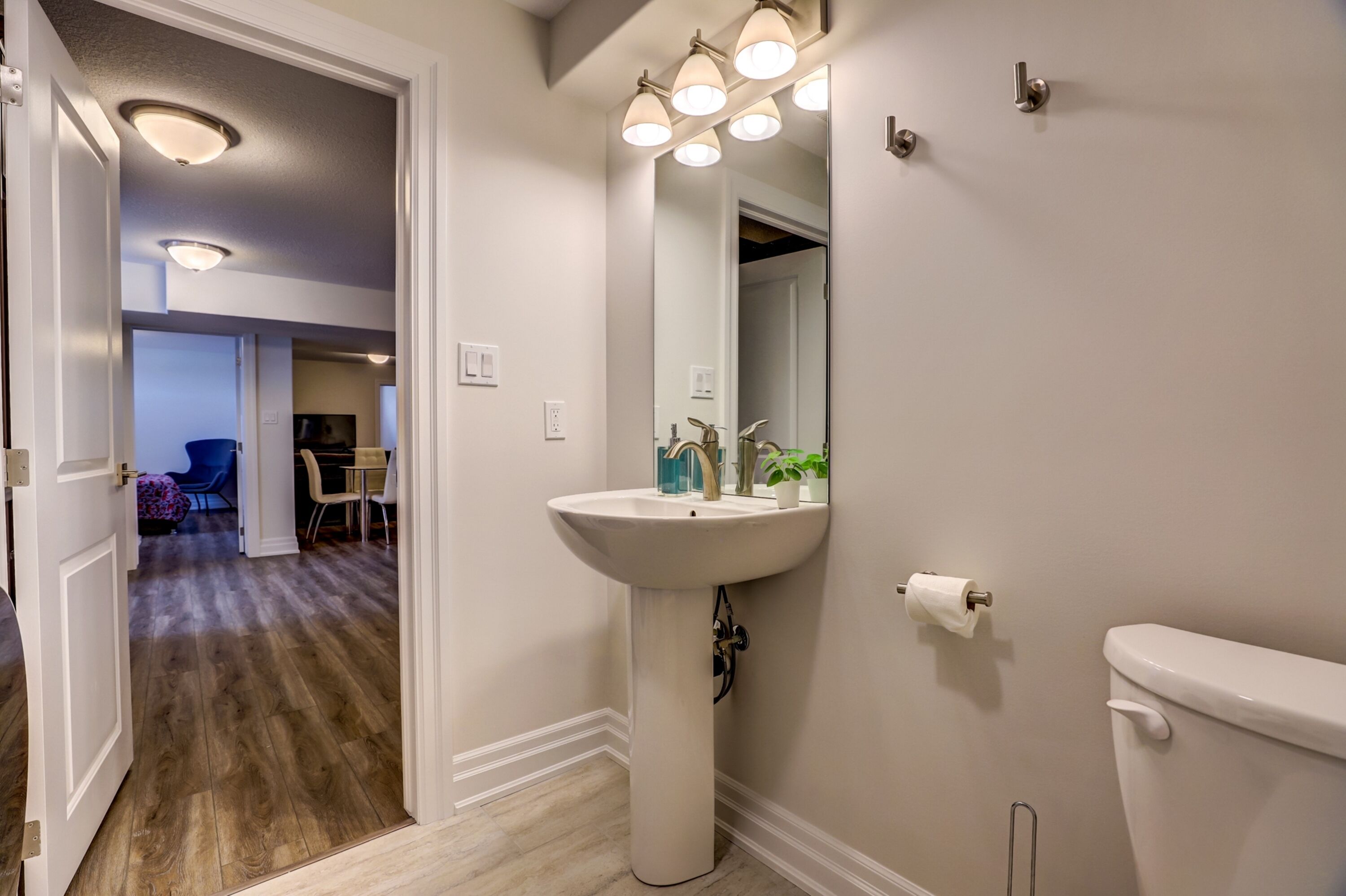 Un bagno con un lavabo su piedistallo, un water e uno specchio illuminato. Si apre una porta che conduce in un'altra stanza.