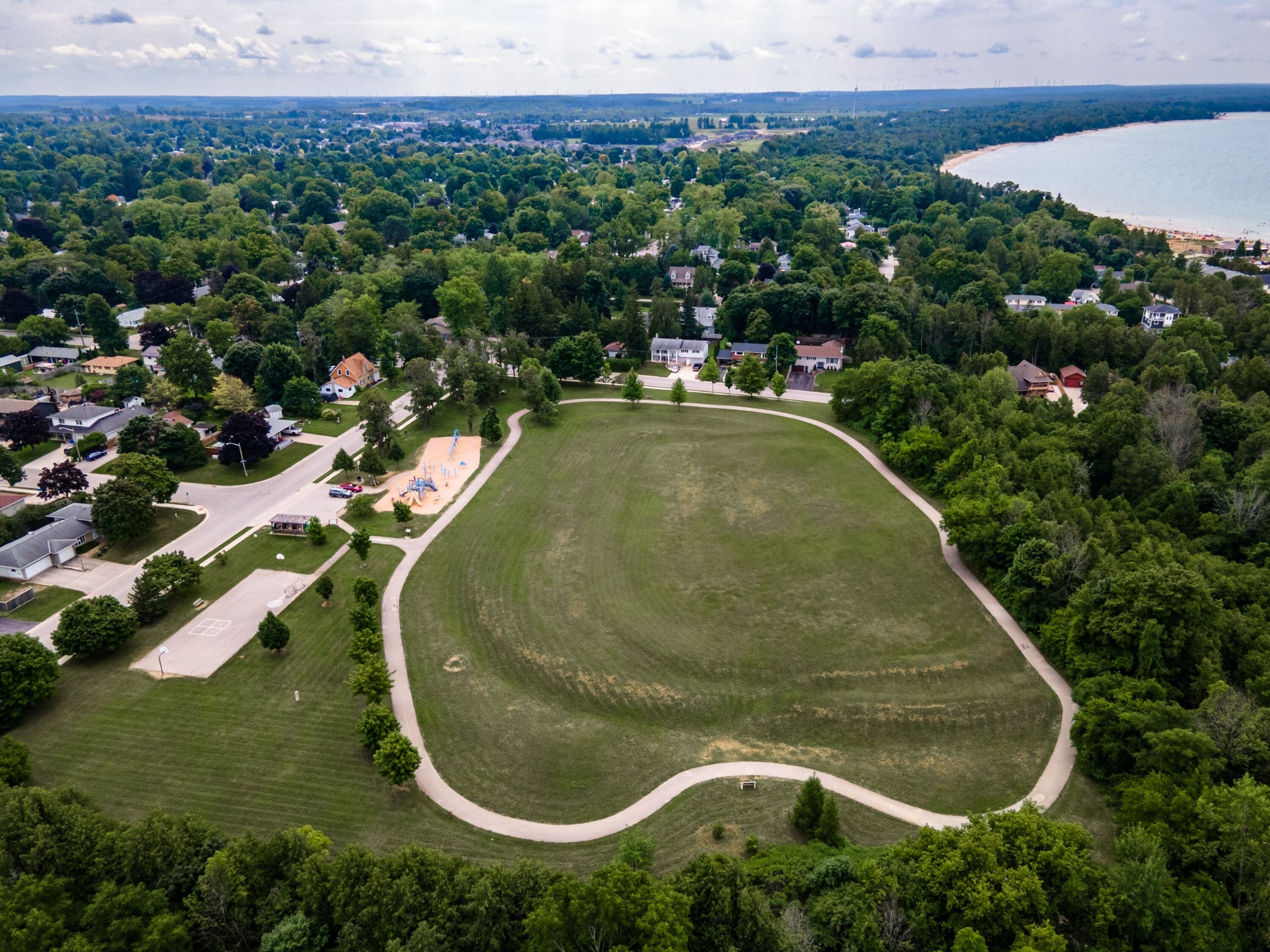 An aerial view of a residential area with a large green park, houses, and a beach in the background.