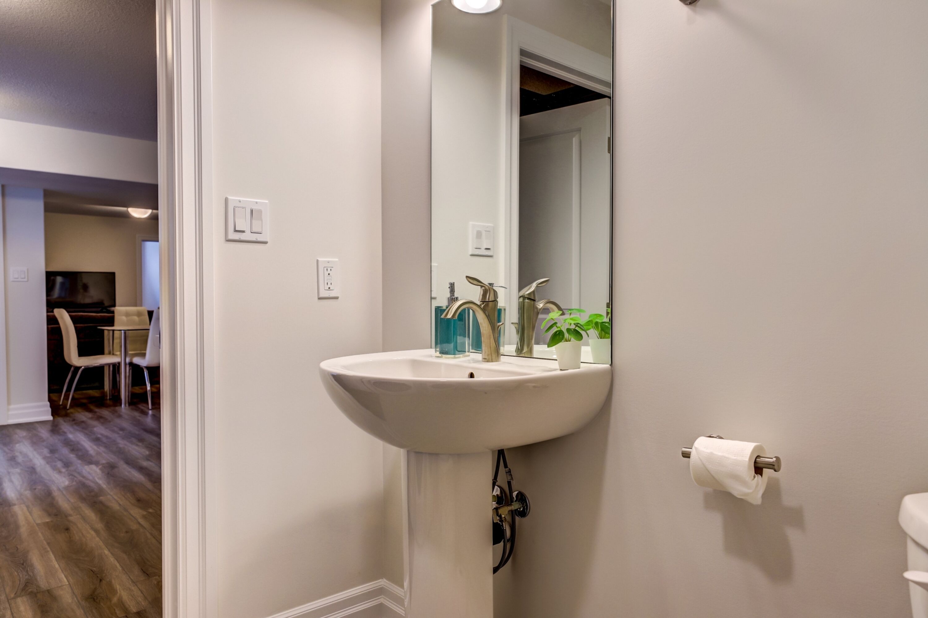 A bathroom with a white sink, a mirror, and a toilet. There is a plant on the sink and a chair in the background.