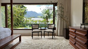 A bedroom with a large window, two chairs, a bed, a wooden dresser, and a view of greenery and mountains. - Vossa Bossa Maresias (São Sebastião)