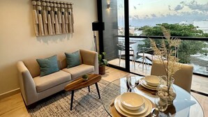 Un salon moderne avec un canapé, une table basse et une table à manger en verre pour deux personnes. Une grande fenêtre offre une vue sur le paysage urbain.