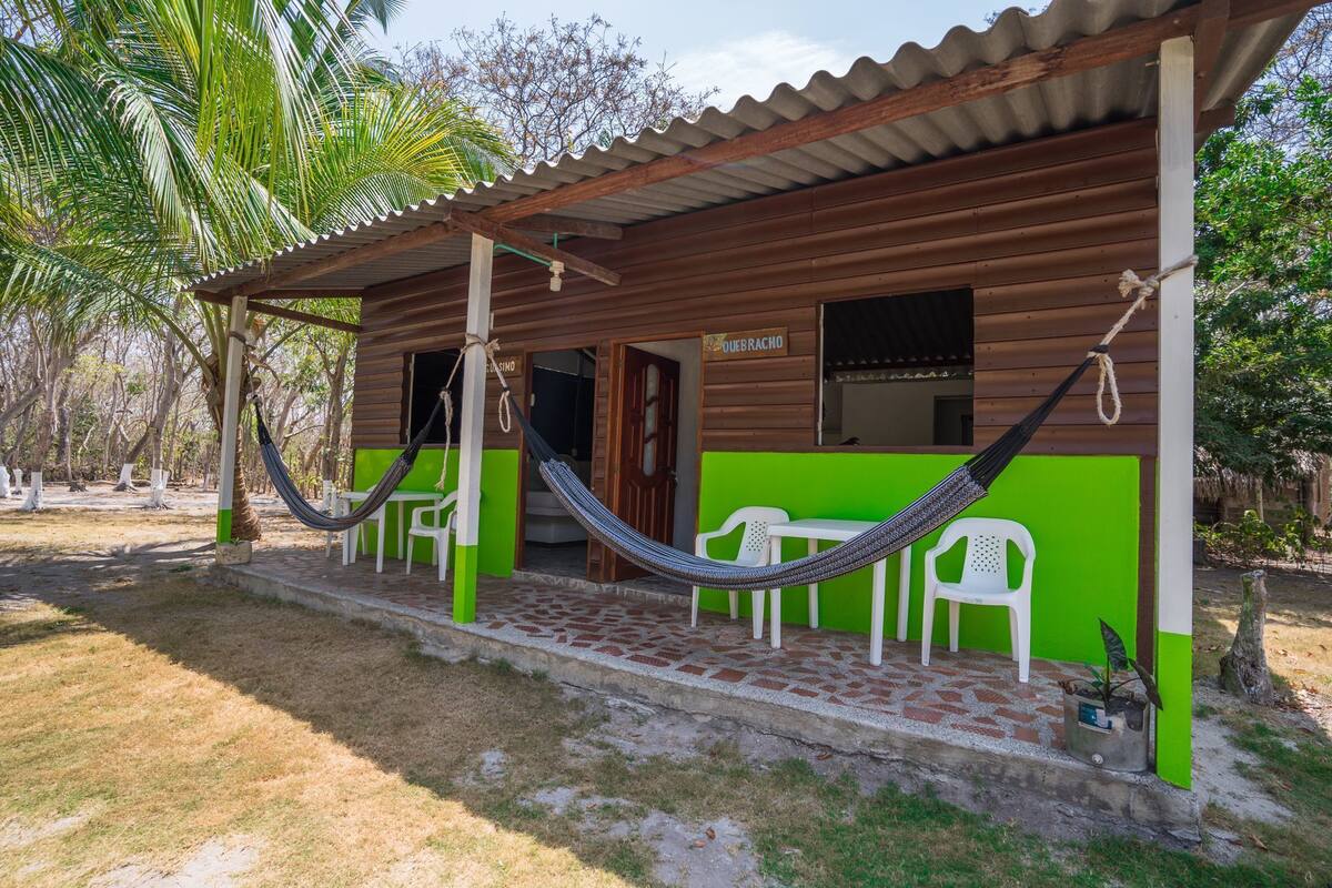 A wooden building with a green facade, a hammock, and a sign that reads "QUEBRADO".