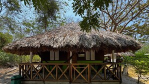 A thatched-roof hut with a wooden railing, situated on sandy ground with trees in the background. - Eco Hotel bosque encantado (Rosario Islands)
