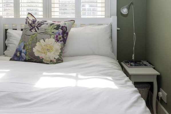 Una cama bien hecha con ropa blanca, un cojín floral, una lámpara de noche y una revista sobre una mesita junto a la cama.