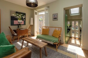 A cozy living room with a sofa, a wooden coffee table, and a dining table. There is a window with a view of the outside.