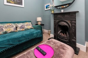 Una sala de estar con un sofá azul aguamarina, una chimenea y un control remoto sobre un pouf rosa.