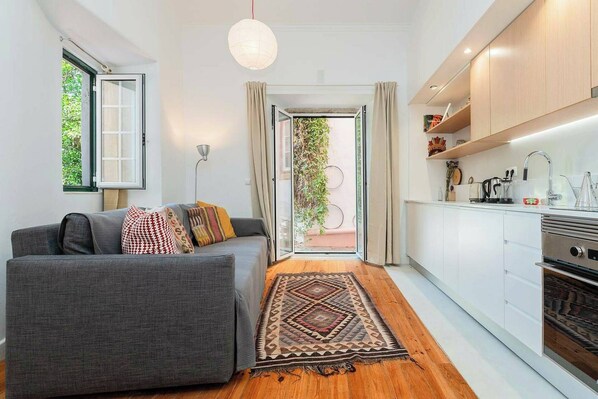 Un salon moderne avec un canapé gris, un tapis à motifs et une cuisine dotée de meubles blancs et d’une cuisinière.