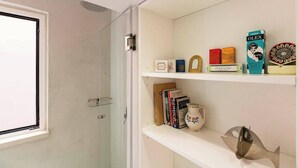 Een badkamer met een douchecabine van glas, een wit schap met boeken en decoratieve objecten, en een raam met uitzicht.