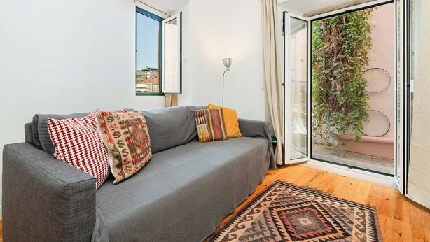 A living room with a grey sofa, patterned cushions, a wooden floor, a view of greenery through a sliding door, and a ceiling light. - Cosy 2 bed flat with terrace near Av Liberdade (Lisboa)