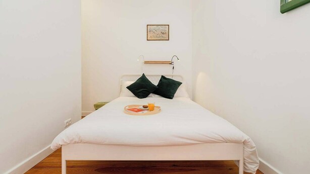 Een kleine kamer met een enkel bed, een nachtkastje, een lijst met een afbeelding aan de muur en een klein krukje.