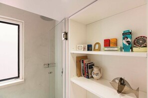 Een badkamer met een douchecabine van glas, een wit schap met boeken en decoratieve objecten, en een raam met uitzicht.