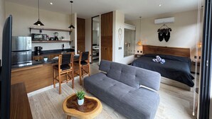 A modern hotel room with a kitchenette, a grey sofa, a wooden coffee table with a plant, and a bed with a dark headboard. - Penthouse Retreat w/Private Rooftop Pool (Tulum)