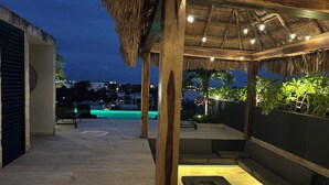 An outdoor seating area with a thatched roof, a fire pit, and a view of a pool and buildings in the background. - Penthouse Retreat w/Private Rooftop Pool (Tulum)