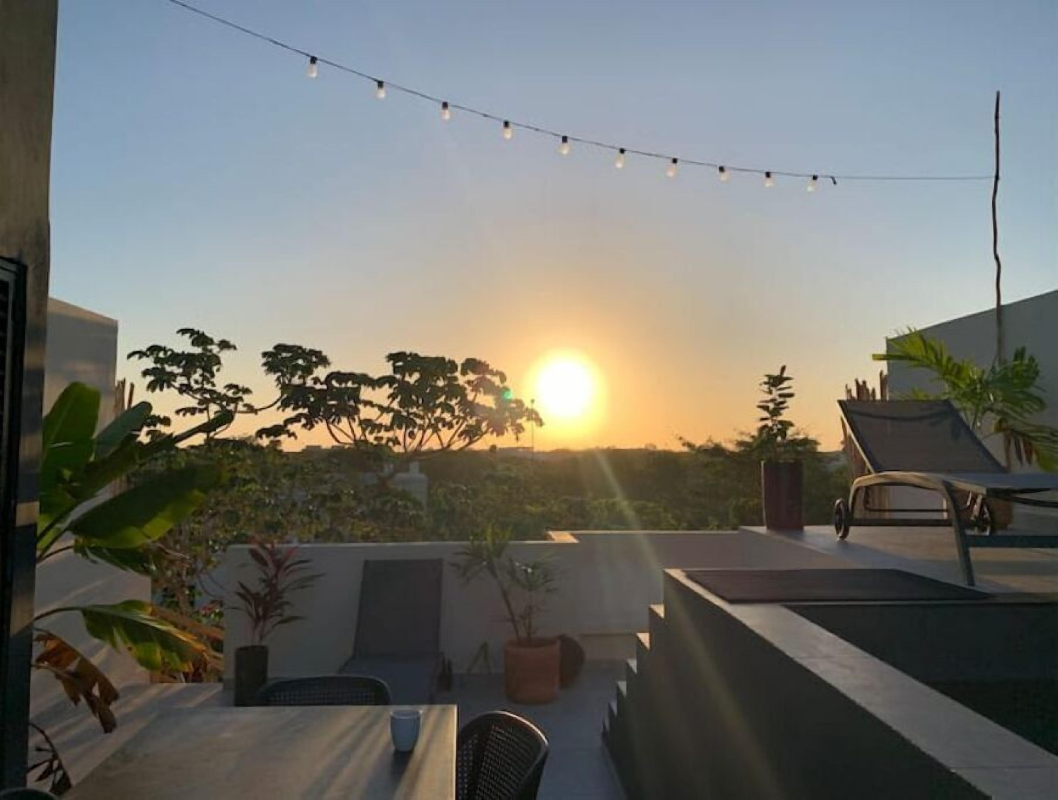 Une terrasse sur le toit, avec une table, des chaises et des guirlandes lumineuses, le tout sur fond de coucher de soleil.