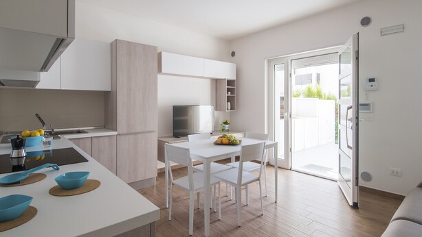 Eine moderne Küche mit weißen Schränken, ein Essbereich mit weißem Tisch und Stühlen sowie eine Glasschiebetür, die auf einen Balkon führt.