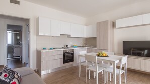 Eine moderne Küche mit weißen Schränken, ein Essbereich mit Tisch und Stühlen sowie ein Fernseher auf der rechten Seite.