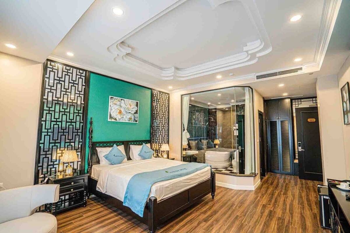A modern hotel room with a large bed, a glass-enclosed bathroom, and a wooden floor.