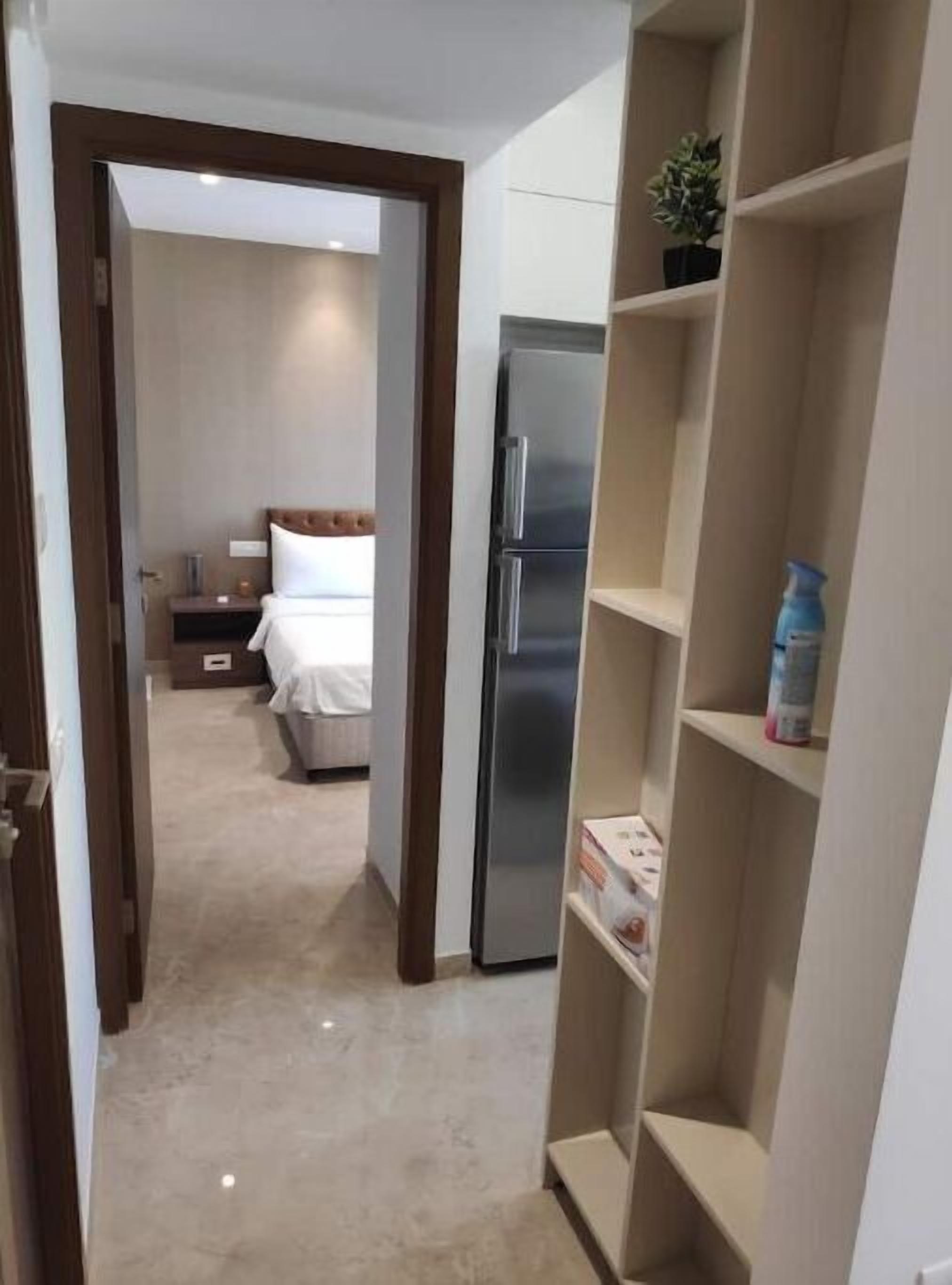 A narrow hallway leading to a bedroom with a bed, a nightstand, and a refrigerator. There is a shelving unit with a potted plant and a spray bottle on it.