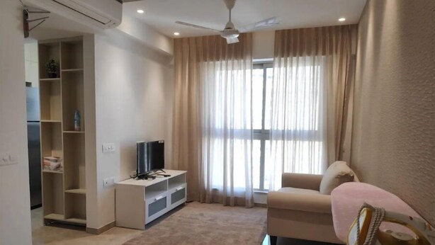 Uma sala de estar moderna com uma TV de tela plana em um suporte branco, cortinas bege, um sofá bege e uma pequena mesa com um ferro de passar roupa, e chão de azulejo.