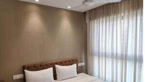 Quarto de hotel com uma cama grande, cabeceira de madeira, roupa de cama branca e uma janela com cortinas leves.