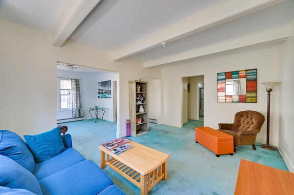Uma sala de estar espaçosa com um sofá azul, um banquinho laranja e uma mesa de centro de madeira. Há uma estante com diversos objetos e um quadro abstrato colorido na parede.
