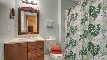 A bathroom with a vanity, a toilet, a shower curtain with a leaf pattern, and a mirror with lights above it.