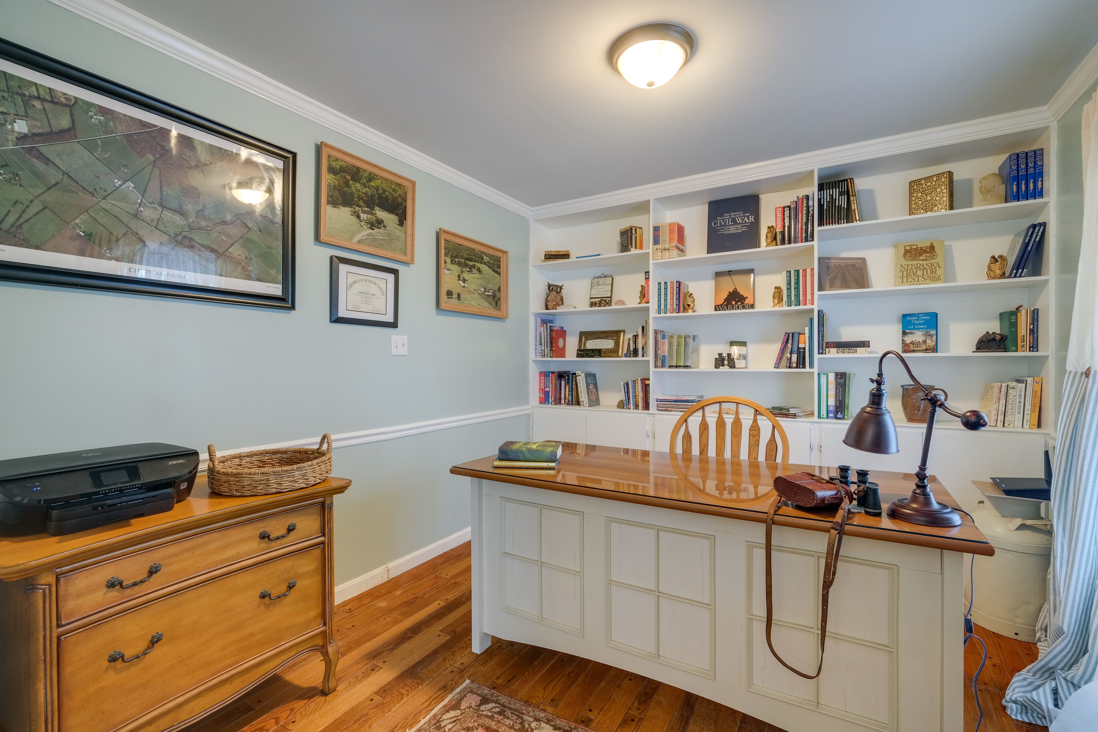 Un estudio con un escritorio de madera, estanterías, una impresora y cuadros en la pared.
