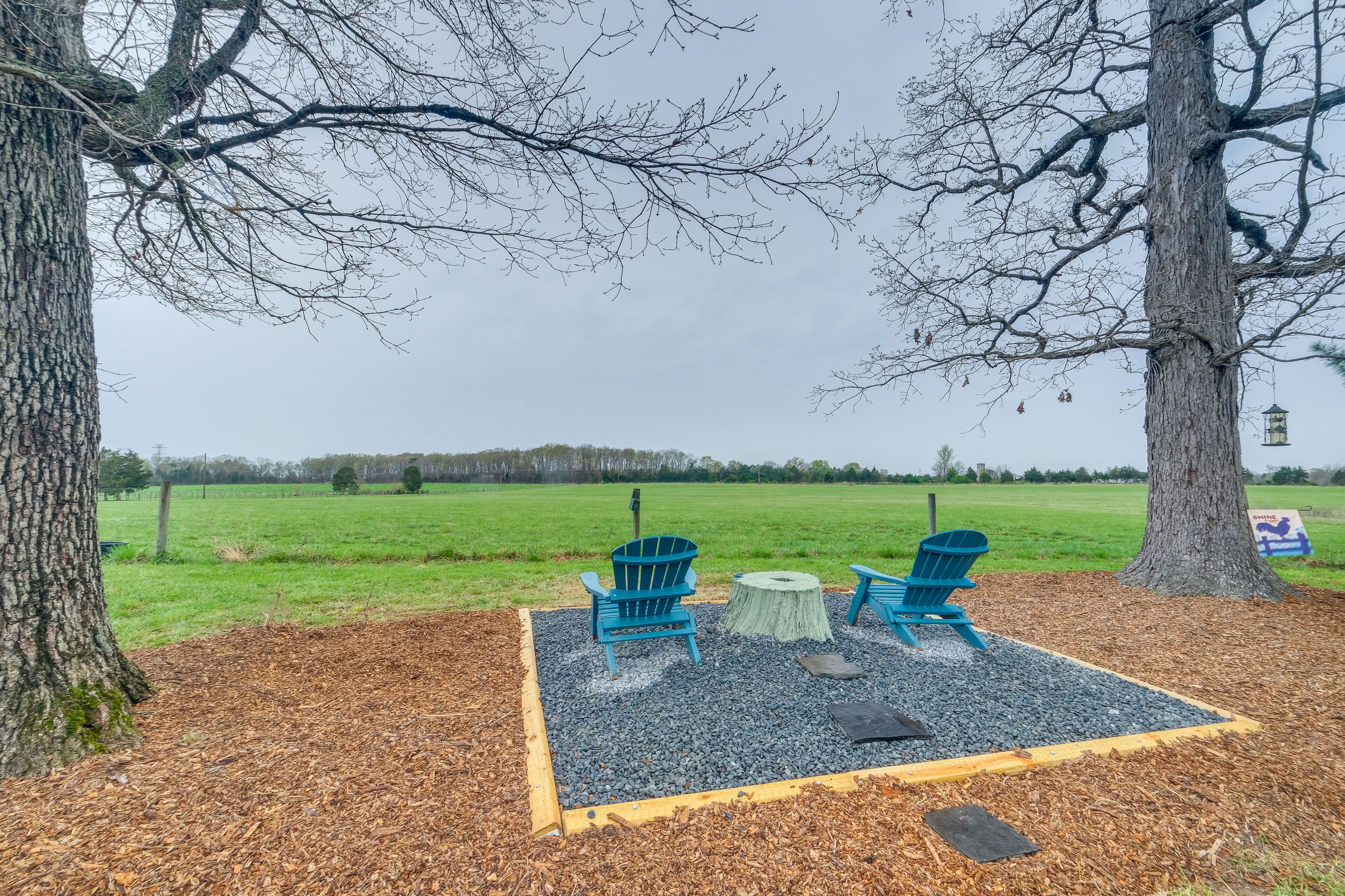 A seating area with two chairs and a small table, surrounded by mulch and gravel, with trees and a grassy field in the background.
