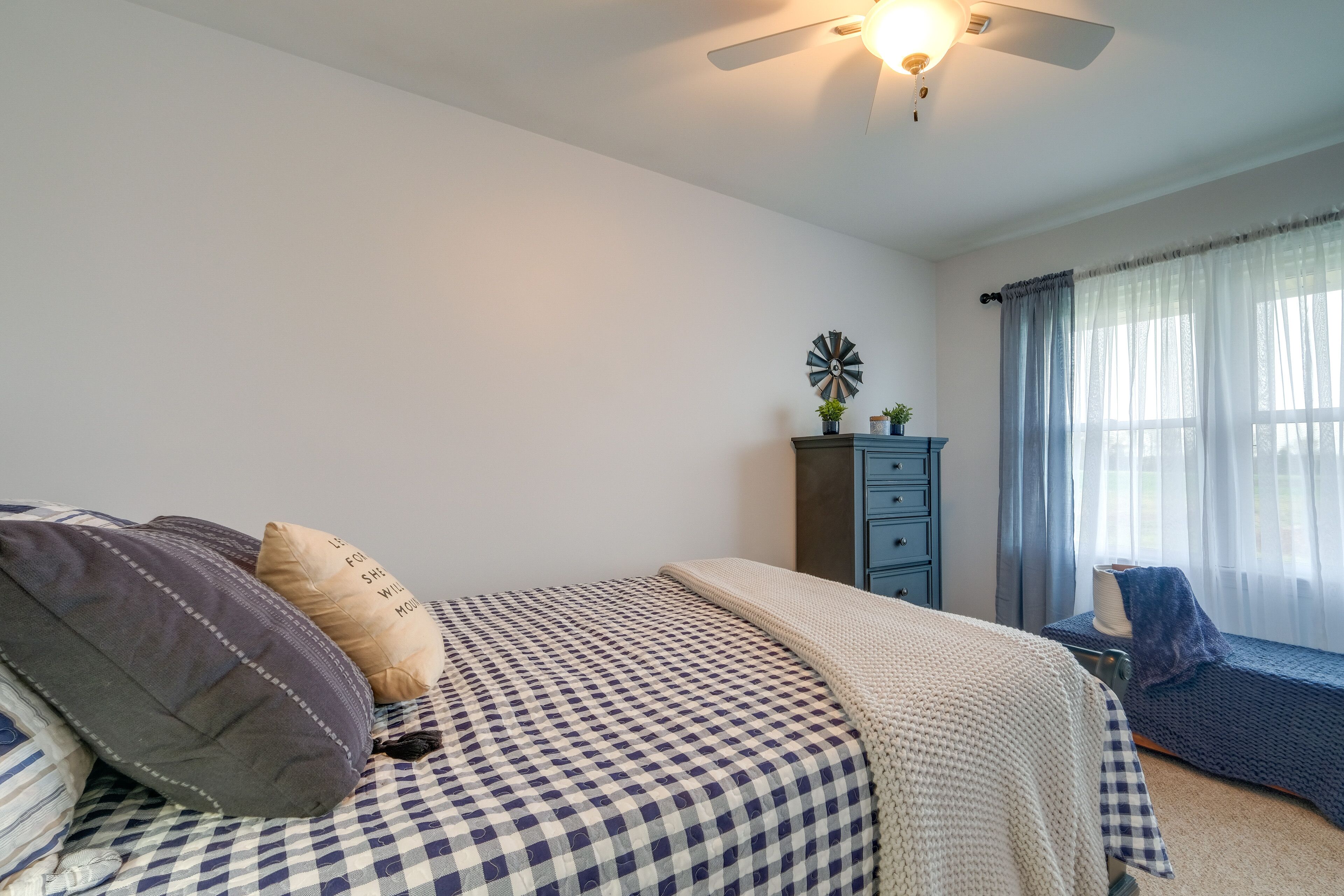 Ein Schlafzimmer mit kariertem Bettbezug, Deckenventilator, Kommode und Fenster mit durchsichtigen Vorhängen.