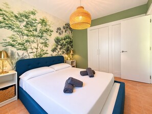 A bedroom with a bed, a nightstand, a wall-mounted lamp, a wardrobe, and a mural of trees.