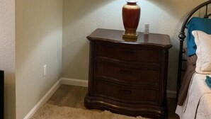 Een slaapkamer met een schuine kap, een houten nachtkastje met een lamp, een bed met een blauwe deken en een beige vloerkleed.