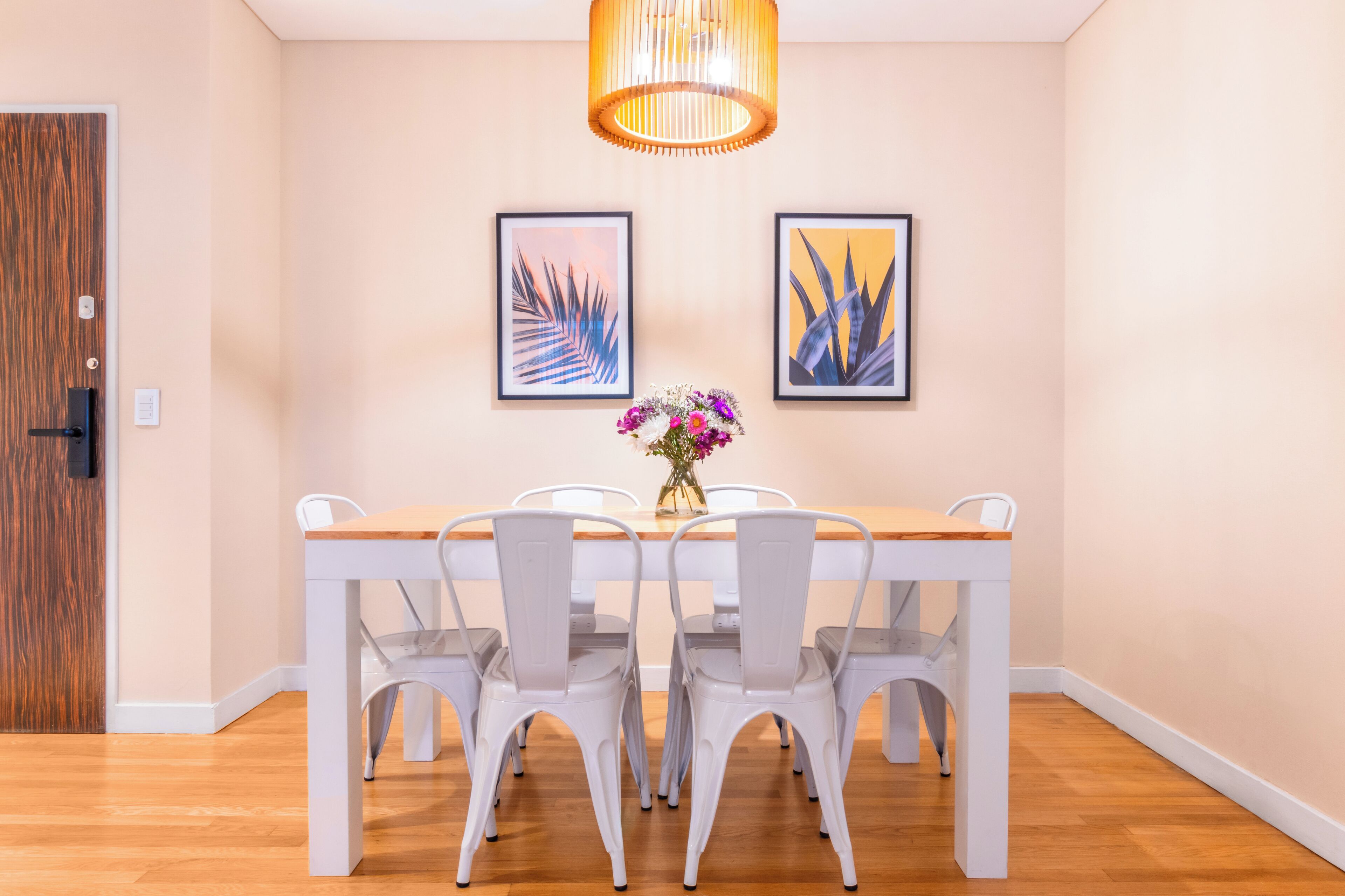 A dining area with a wooden table and white chairs, framed artwork on the wall, and a hanging light fixture.