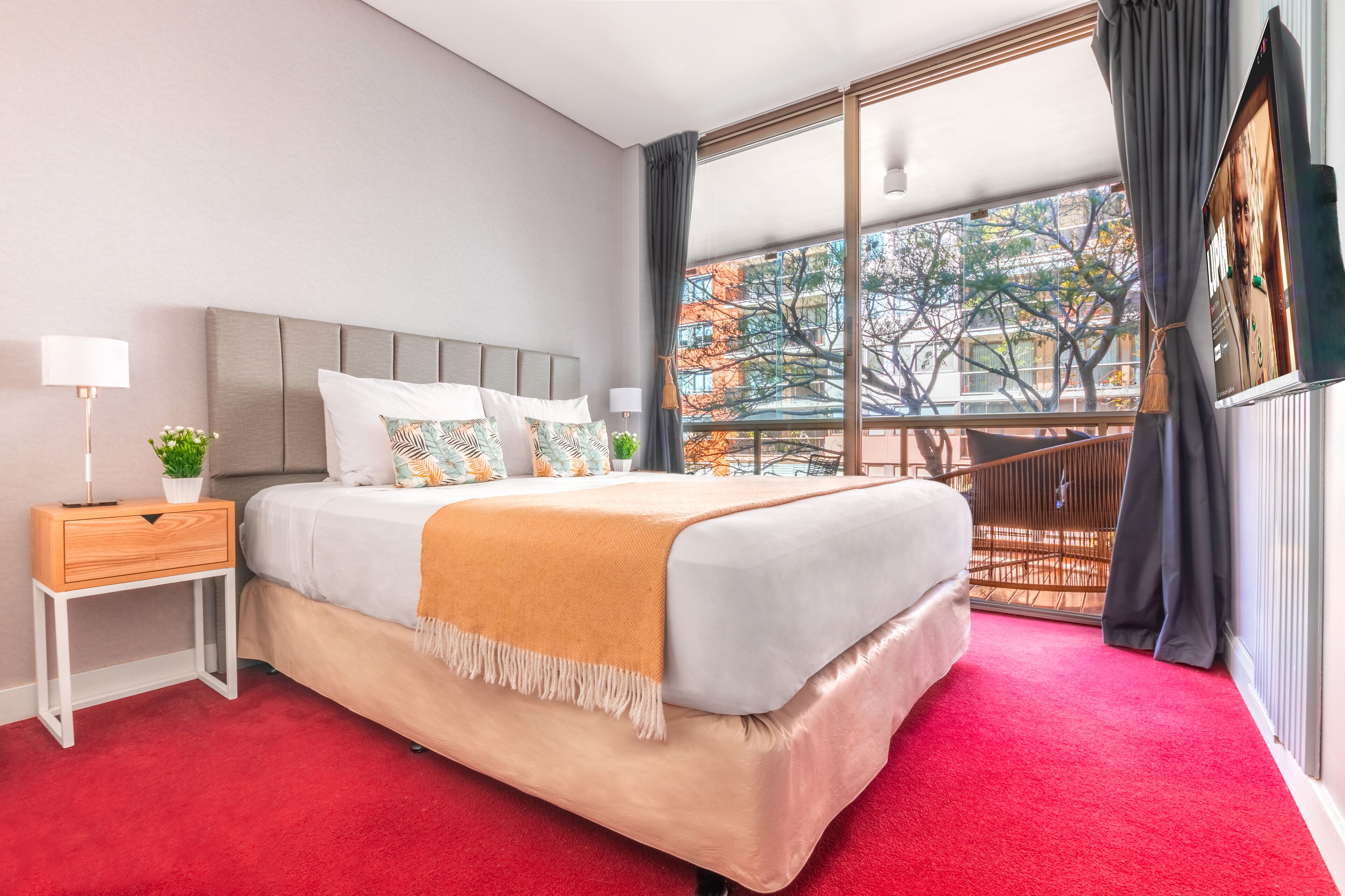 A modern hotel room with a large bed, a nightstand, a window with a view of trees, and a balcony.
