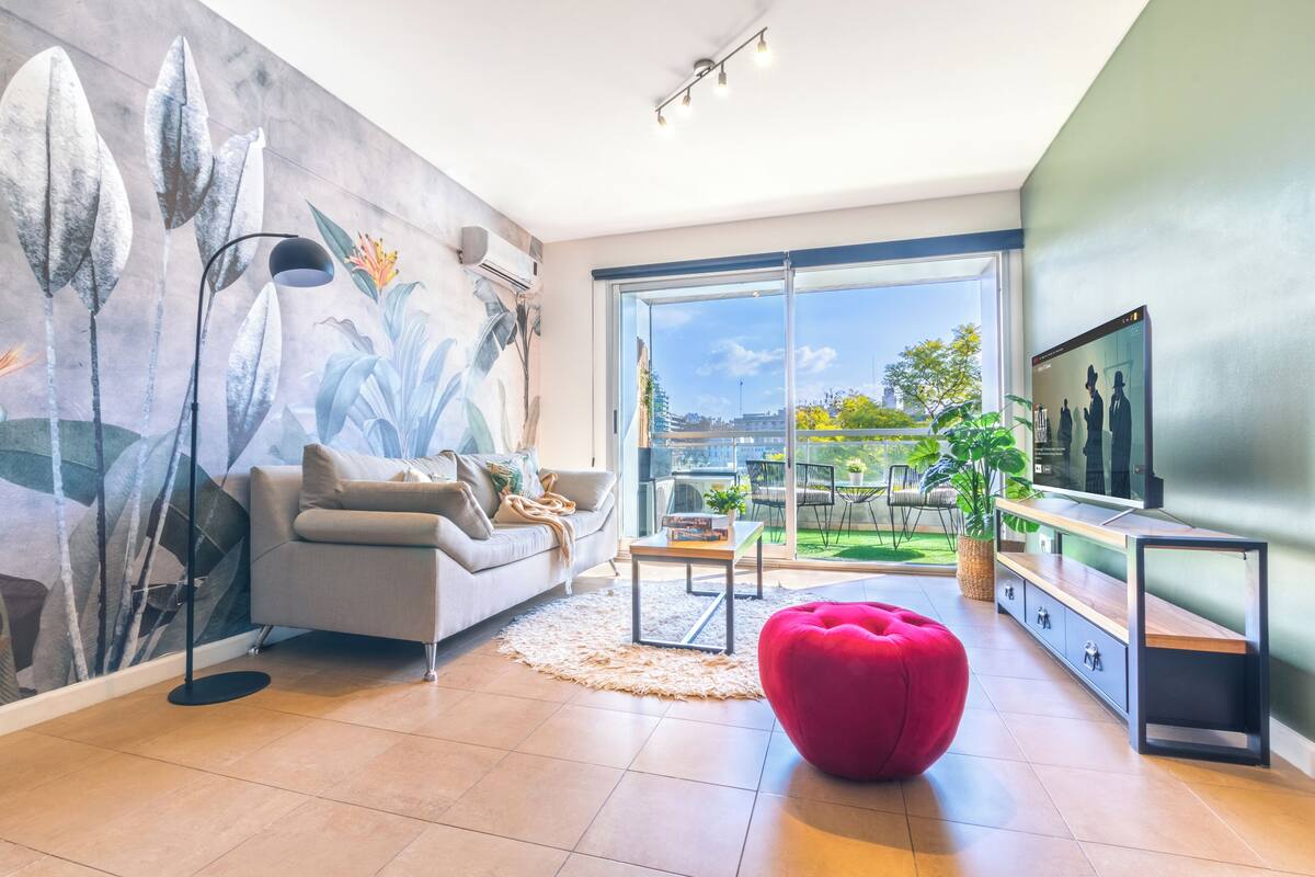 Una sala de estar moderna con un sofá gris, un reposapiés rojo, un mural grande, un televisor y un ventanal corredizo que da a un área exterior.