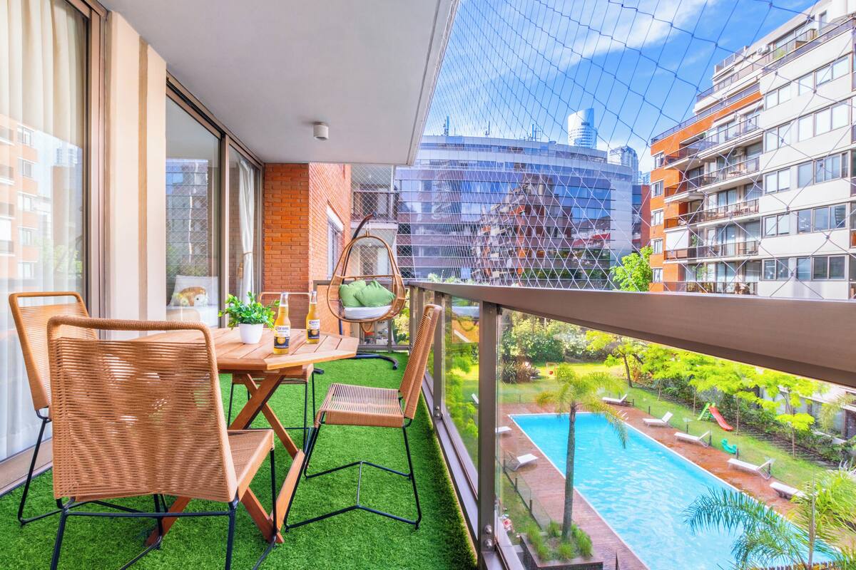 Un balcón con vista a una piscina, una mesa de comedor con sillas y un ventanal que da a una habitación.