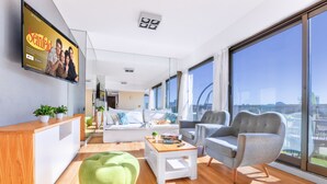 Ein modernes Wohnzimmer mit großem Fernsehbildschirm, einem Holzbüro, einem grünen Sitzsack, zwei grauen Sesseln und einem weißen Sofa.