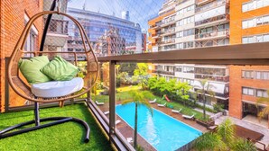 Ein Balkon mit Korbstuhl, Blick auf einen Swimmingpool und umgebende Gebäude.