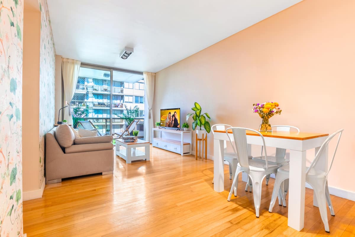 Una sala moderna con sofá, mesa de centro, mesa de comedor con sillas y vistas a un edificio a través de amplios ventanales.