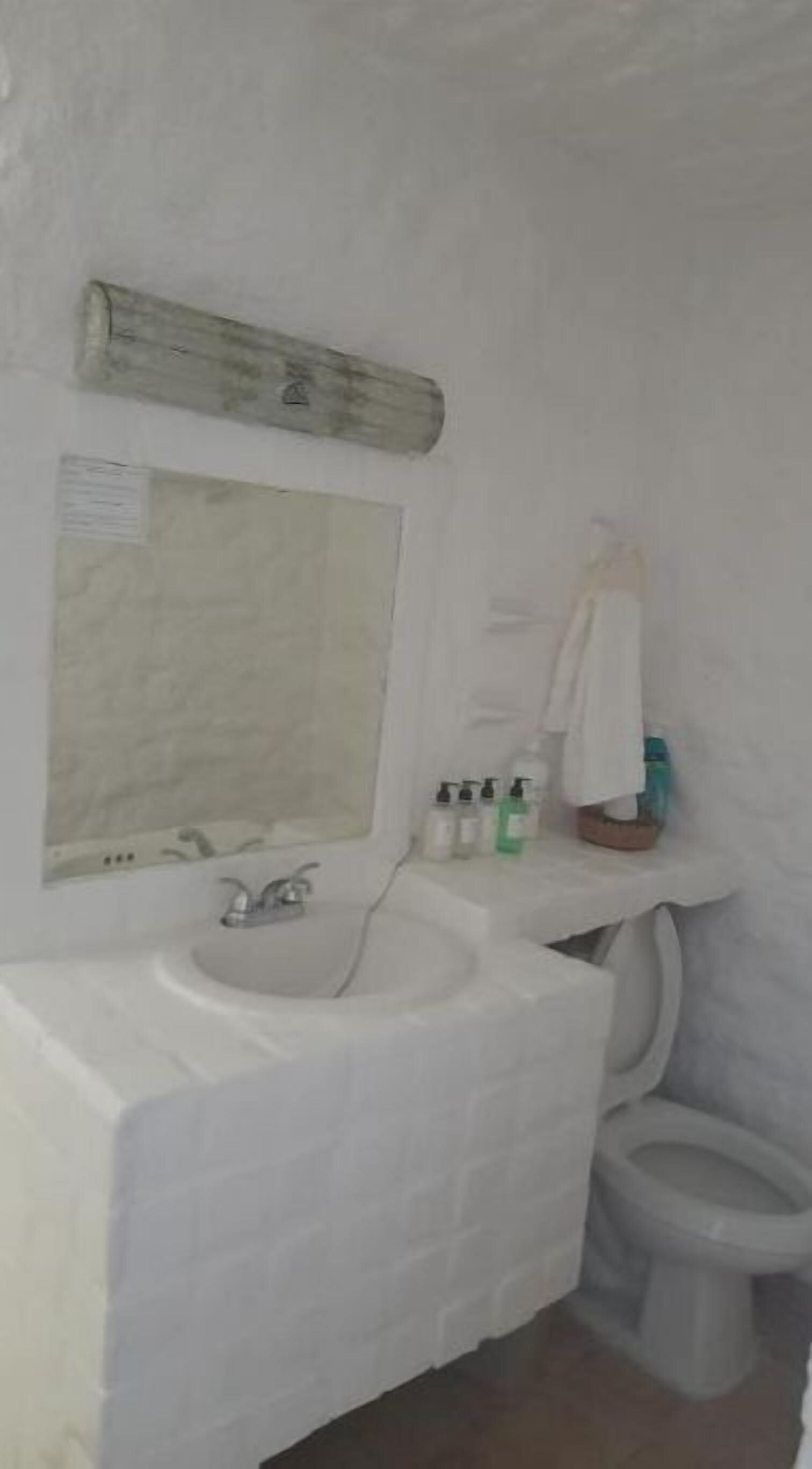 Un baño con un lavabo blanco, un espejo y un inodoro. Hay artículos de aseo en el lavabo y una toalla colgada en la pared.