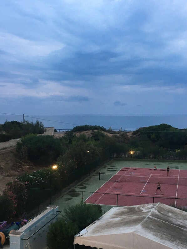 Pista de tenis con red, rodeada de vegetación y vistas al mar.